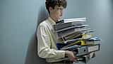 An office worker carries a massive, heavy stack of colourful document folders in a modern workplace.