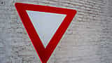 A red and white triangular yield sign stands in front of a white brick wall.