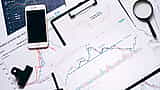 A top-down view of a desk displaying a smartphone, magnifying glass, and printed financial charts analyzing S&P 500 and Nasdaq market returns.