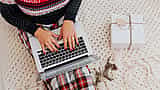 A person wearing a festive sweater and plaid pants types on a laptop next to a wrapped holiday gift on a knitted blanket.