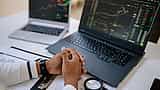 A person sits at a desk with folded hands, monitoring financial charts on two laptop screens.