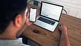A person sits at a wooden desk analyzing financial market charts simultaneously on a smartphone and a laptop.