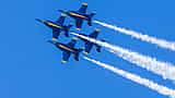 Four U.S. Navy Blue Angels F/A-18 fighter jets soar in tight formation with white smoke trails against a bright blue sky.