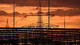 Silhouetted electrical transmission towers stand against a vibrant orange and yellow sunset.