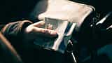 A person's hand holding a blue Canadian five-dollar bill next to a leather handbag.