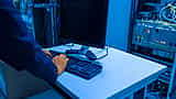 A person's hands type on a computer keyboard at a workstation inside a blue-lit server room.