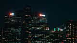 Illuminated logos of major banks shine from skyscrapers against a dark night sky.