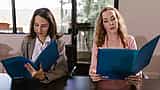 Two professional women sit side-by-side at a conference table, each reading from identical blue folders.