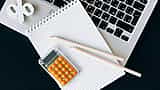 A top-down view of a modern desk featuring a laptop, grid notebook, orange calculator, and a white percent symbol.