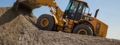 A yellow Caterpillar wheel loader moves a large mound of gravel against a clear blue sky.