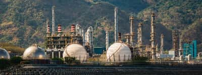 A large industrial oil refinery with spherical storage tanks and tall towers sits near a body of water.