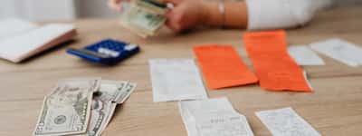 A person uses a calculator next to a small stack of cash on a wooden desk.