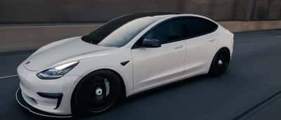 A white Tesla Model 3 driving swiftly along a highway.