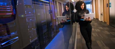A professional holds a laptop next to glass-enclosed server racks in a modern data center.