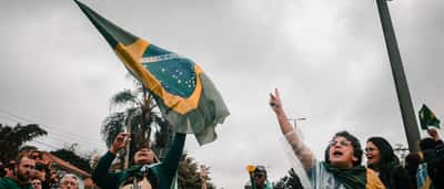 A crowd of passionate demonstrators, with a woman in the foreground cheering while raising the Brazilian flag under a cloudy sky.