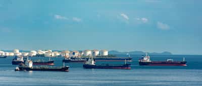 Several oil tankers wait on the water in front of a coastal fuel storage terminal with large white tanks.