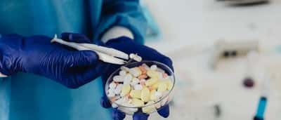 Gloved hands use tweezers to carefully examine a collection of pills in a clear glass petri dish.