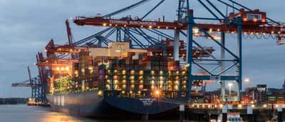 A large cargo ship loaded with shipping containers is docked beneath towering cranes at a port terminal in Hamburg, Germany.
