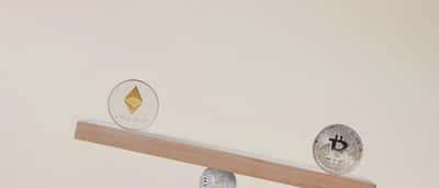 A silver Ethereum coin and a silver Bitcoin coin balancing on opposite ends of a wooden seesaw against a beige background.