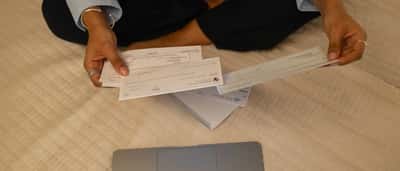 A person's hands hold printed checks over a desk with a laptop.
