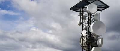 A tall telecommunications tower equipped with large white satellite dishes stands against a cloudy sky.