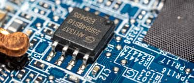 A black microchip is soldered onto a blue printed circuit board among other small electronic components.