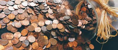 A glass jar lies on its side, spilling a large mound of copper pennies onto a flat surface.