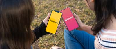 Two people sit outdoors holding smartphones that display the Bumble and Tinder dating apps.