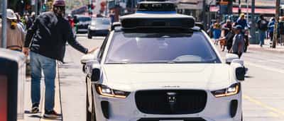 A white Waymo self-driving vehicle with roof-mounted sensors navigates a city street.