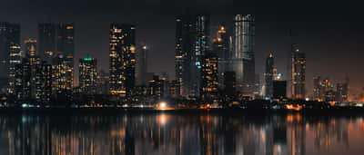 The modern high-rises of the Mumbai skyline glow at night, reflecting off the dark waters below.