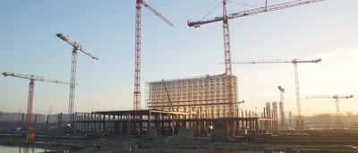 Multiple towering cranes stand over a massive building framework at a large-scale construction site at dusk.