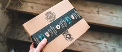 A hand holds a cardboard delivery box sealed with Amazon Prime tape over wooden steps.