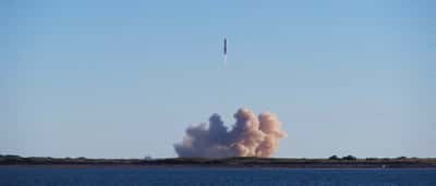 A rocket launches into a clear blue sky, leaving a thick trail of white smoke.