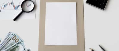 A blank piece of white paper sits on a desk surrounded by stock charts, a magnifying glass, a calculator, and hundred-dollar bills.