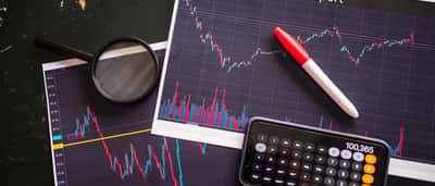A printed stock report showing candlestick charts, resting on a dark surface alongside a magnifying glass, marker, and a calculator on a smartphone.