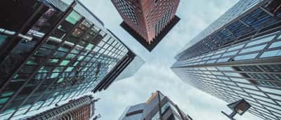A low-angle view looking up at towering glass skyscrapers reaching toward a cloudy blue sky.