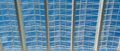 A modern glass ceiling structure looking up toward a bright blue sky.