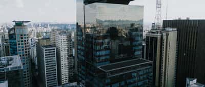 A tall, modern glass skyscraper reflects the dense urban skyline of São Paulo.