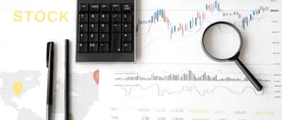 A flat lay of a desk featuring financial line charts, a dotted world map graphic, a calculator, and a magnifying glass.