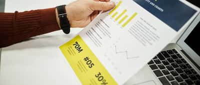 A professional holding a detailed finance report with bar charts over a laptop on a white desk.