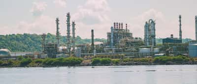 A large industrial oil refinery facility sits by the water under a bright blue, cloudy sky.