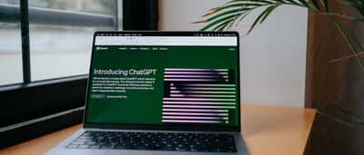 A laptop sits on a wooden desk by a window, displaying the OpenAI website and an introduction to ChatGPT on its screen.