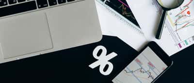 A white percentage symbol rests on a dark desk surrounded by a laptop, a smartphone displaying market data, and printed financial charts.