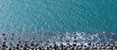 An aerial view of geometric concrete breakwater blocks protecting a shoreline from deep blue ocean waves.