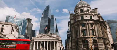 A red double-decker bus passes historic classical buildings and modern glass skyscrapers in the City of London's financial district.