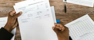 A person's hands hold up a financial statement over a wooden desk with a tax form resting nearby.