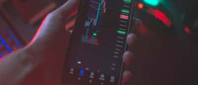 A person's hand holds a smartphone displaying a dark-mode cryptocurrency trading chart with colorful price candles.