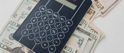A black digital calculator rests on top of a scattered pile of U.S. dollar bills.