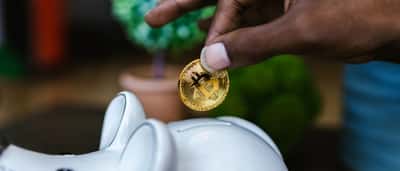 A hand places a gold physical Bitcoin token into the slot of a white ceramic piggy bank.