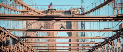 Looking through the intricate steel beams of the Brooklyn Bridge toward its stone arches and an American flag.